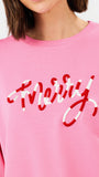 A woman wearing a pink crewneck sweatshirt with red script embroidery spelling 'merry' on the front, paired with blue jeans, against a plain white studio background.