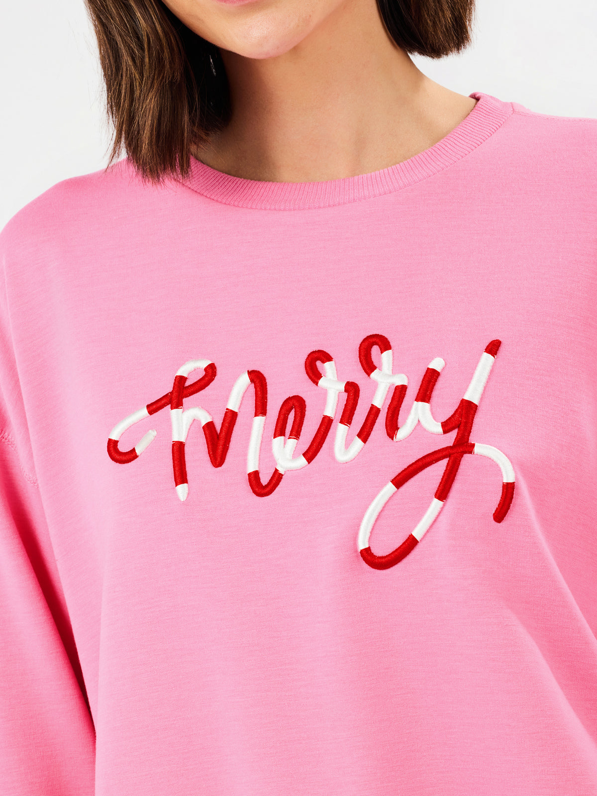 A woman wearing a pink crewneck sweatshirt with red script embroidery spelling 'merry' on the front, paired with blue jeans, against a plain white studio background.