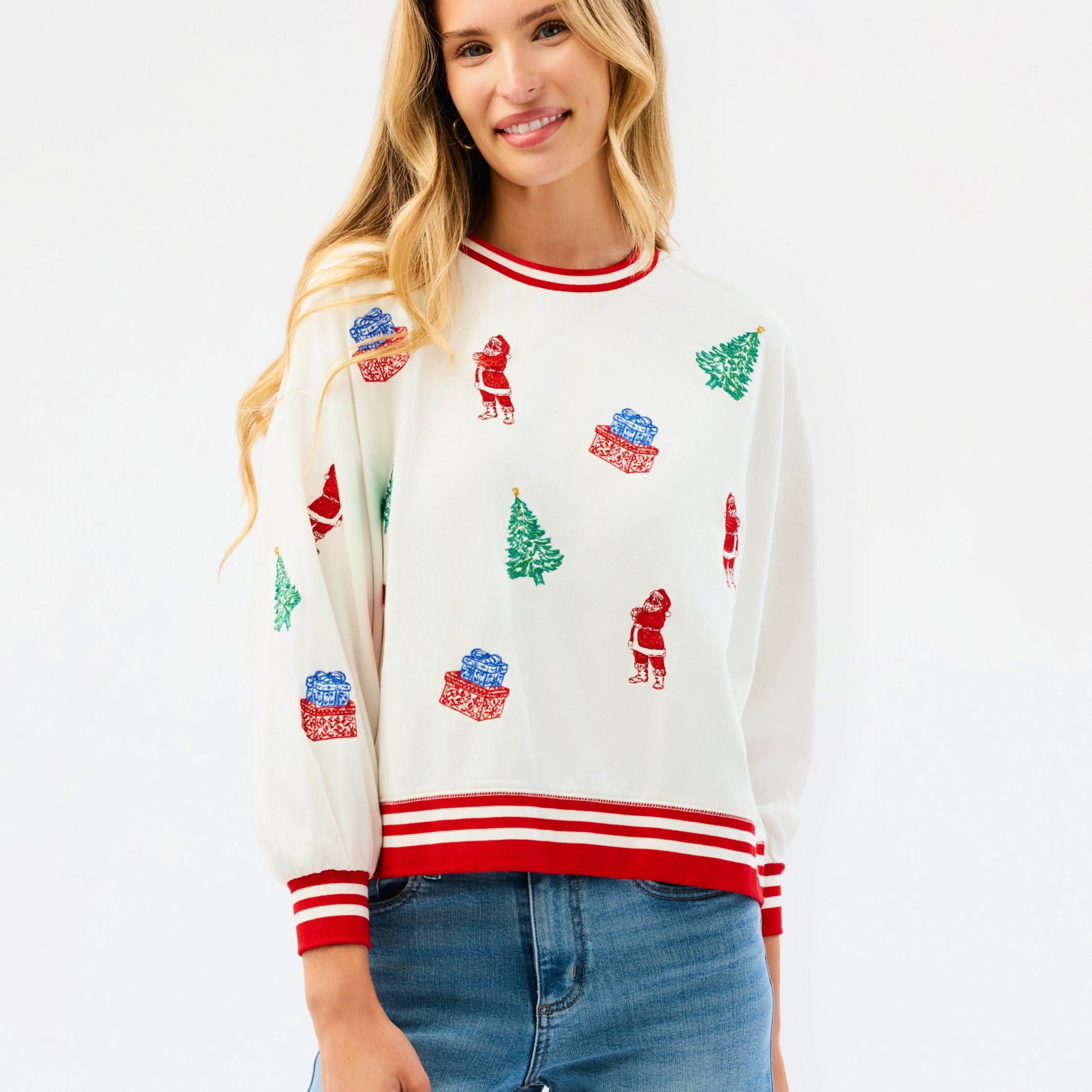 A woman wearing a cream-colored Christmas crewneck sweater with red and white striped trim and green/red holiday motifs, paired with blue jeans, against a plain white background.