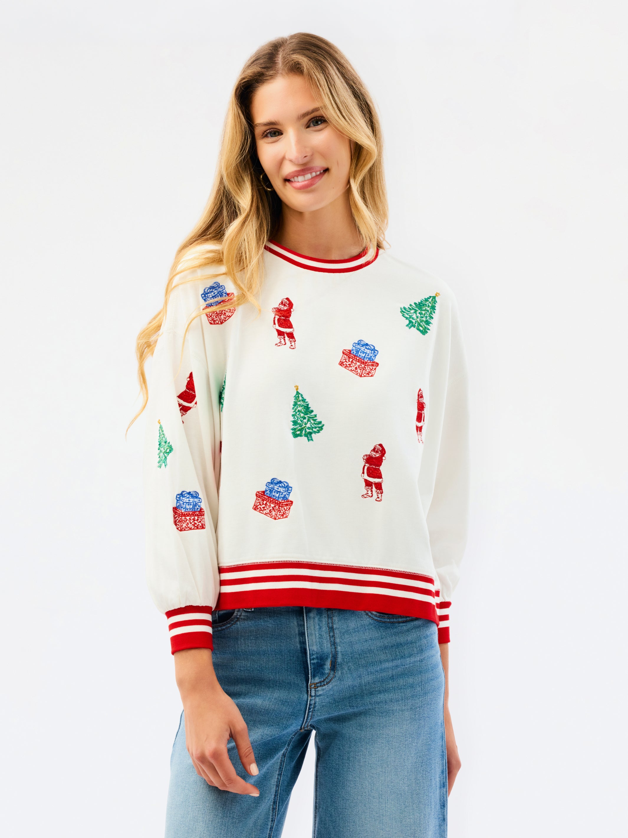 A woman wearing a cream-colored Christmas crewneck sweater with red and white striped trim and green/red holiday motifs, paired with blue jeans, against a plain white background.