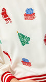 Close-up of a cream-colored knit sweater with a festive Christmas print featuring red Santa Claus figures, green Christmas trees, and red and blue gift boxes, finished with red and white striped ribbed cuffs and hem.