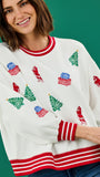 A woman wearing a white crewneck sweatshirt decorated with red and green Christmas-themed graphics, including Santa Claus figures, Christmas trees, and wrapped gifts, set against a solid green background.