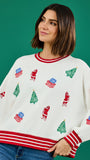A woman wearing a white Christmas-themed crewneck sweater with red striped trim and red, green, and blue holiday motifs, paired with blue jeans against a solid green background.