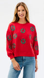 A woman modeling a red crewneck sweatshirt decorated with green Christmas wreaths, paired with blue jeans, against a plain white studio background.