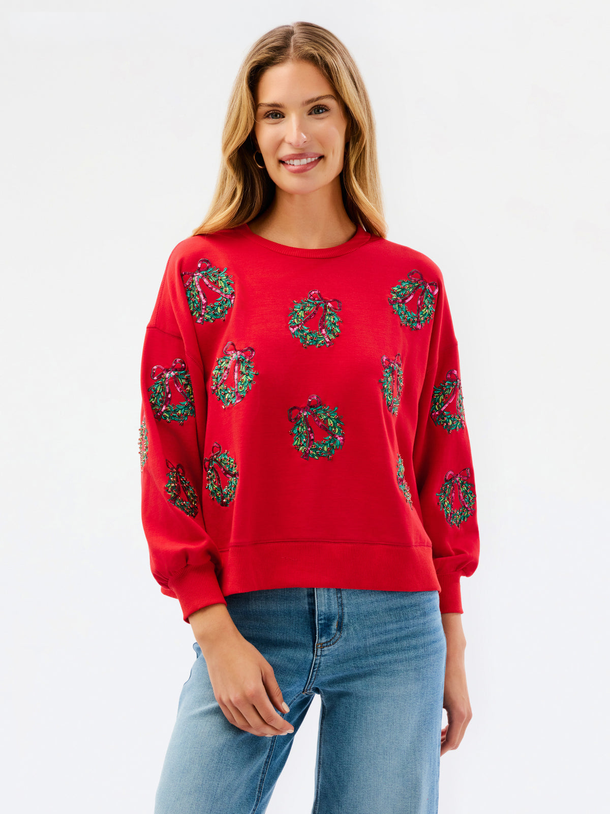 A woman modeling a red crewneck sweatshirt decorated with green Christmas wreaths, paired with blue jeans, against a plain white studio background.