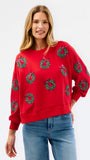 A woman wearing a bright red crewneck sweatshirt decorated with green and black Christmas wreath patterns, paired with blue jeans, against a plain white studio background.