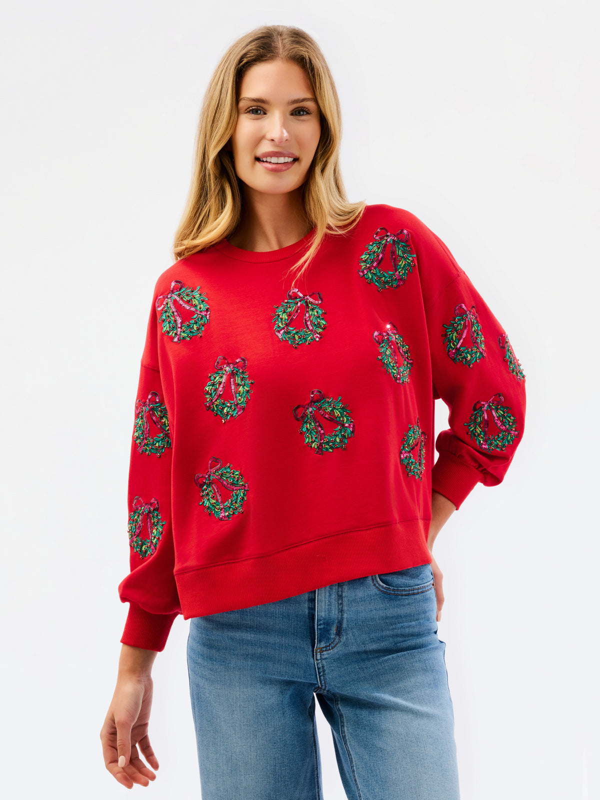 A woman wearing a bright red crewneck sweatshirt decorated with green and black Christmas wreath patterns, paired with blue jeans, against a plain white studio background.