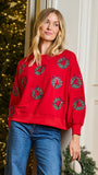 A woman wearing a festive red crewneck sweatshirt with green wreath patterns, paired with blue jeans, standing in front of a decorated Christmas tree and a white paneled wall.