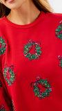 A close-up of a bright red crewneck sweatshirt featuring green and pink sequined Christmas wreath motifs with red bows.