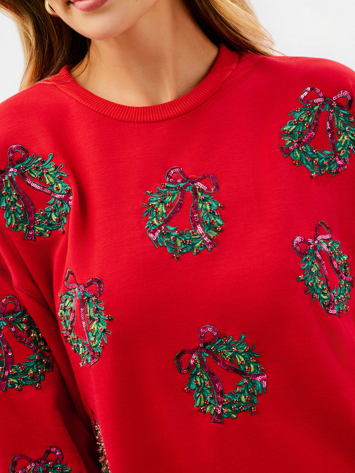 A close-up of a bright red crewneck sweatshirt featuring green and pink sequined Christmas wreath motifs with red bows.