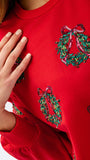 A close-up of a bright red crewneck sweatshirt featuring green holly wreath sequin embellishments with pink bows, worn by a model whose hand is resting on the chest.