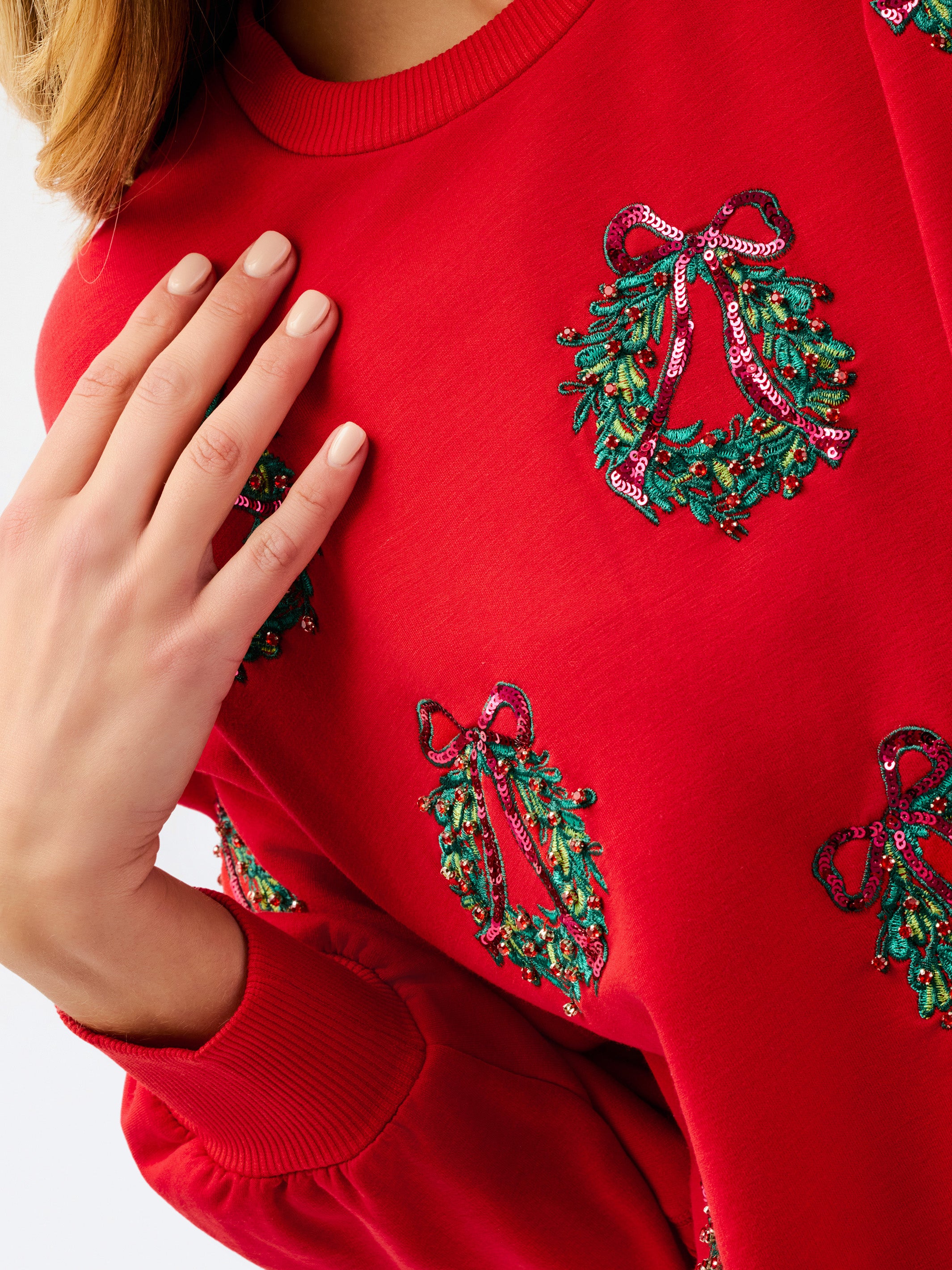 A close-up of a bright red crewneck sweatshirt featuring green holly wreath sequin embellishments with pink bows, worn by a model whose hand is resting on the chest.