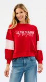 A woman wearing a red crewneck sweatshirt with 'TIS THE SEASON to be jolly' printed in white across the chest and white stripes on the sleeves, paired with blue jeans, against a plain white background.