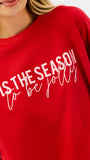 A close-up of a person wearing a bright red crewneck sweatshirt with white text that reads 'TIS THE SEASON to be jolly'.