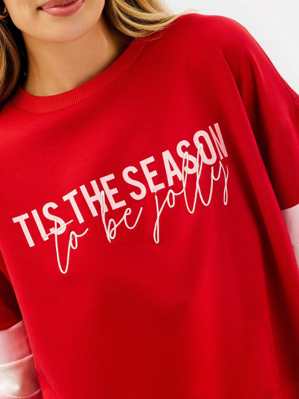 A close-up of a person wearing a bright red crewneck sweatshirt with white text that reads 'TIS THE SEASON to be jolly'.