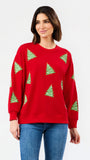 A woman wearing a red crewneck sweatshirt with green Christmas tree patterns, paired with blue jeans, against a plain white studio background.