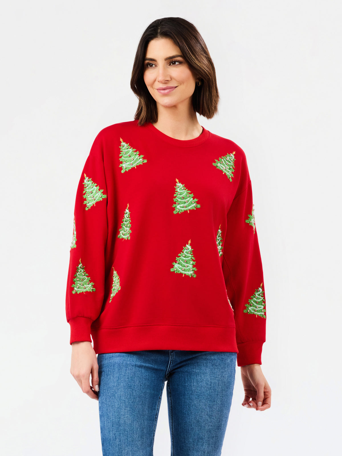A woman wearing a red crewneck sweatshirt with green Christmas tree patterns, paired with blue jeans, against a plain white studio background.