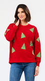 A woman wearing a red crewneck sweatshirt with green Christmas tree patterns, paired with blue jeans, against a plain white studio background.