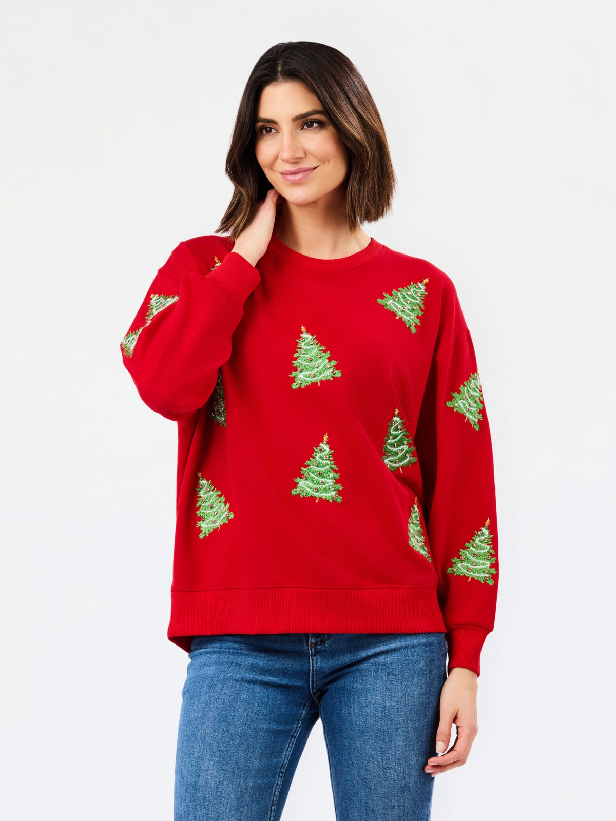 A woman wearing a red crewneck sweatshirt with green Christmas tree patterns, paired with blue jeans, against a plain white studio background.