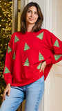 A woman wearing a bright red crewneck sweatshirt with green Christmas tree patterns, paired with light blue jeans, standing in front of a decorated Christmas tree and a cream-colored door.