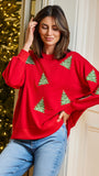 A woman wearing a festive red crewneck sweatshirt with green embroidered Christmas trees, paired with blue jeans, posing in front of a decorated Christmas tree and a cream-colored door.
