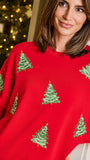 A woman wearing a bright red crewneck sweatshirt decorated with green embroidered Christmas trees, posed in front of a softly blurred festive background with warm lights.