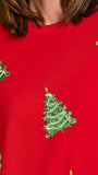A close-up of a bright red crewneck sweater featuring embroidered green Christmas trees with small gold and red accents, worn by a person.