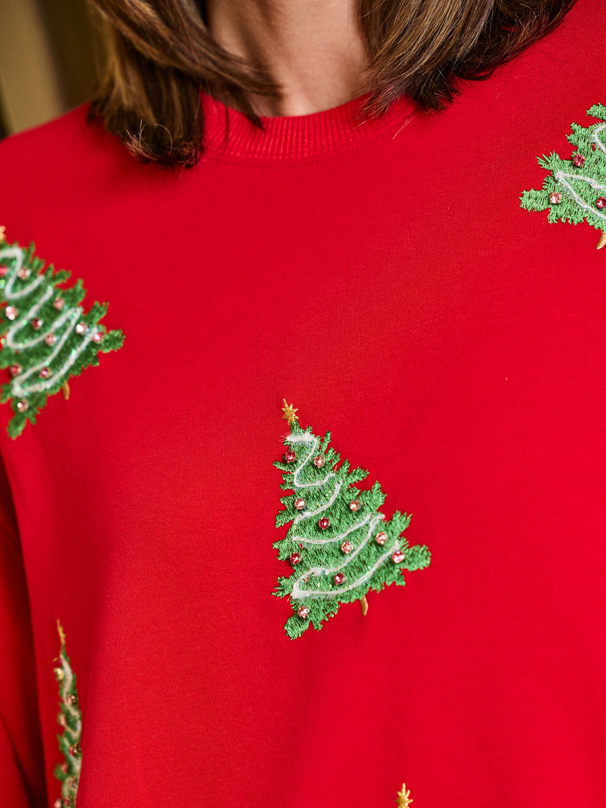 A close-up of a bright red crewneck sweater featuring embroidered green Christmas trees with small gold and red accents, worn by a person.