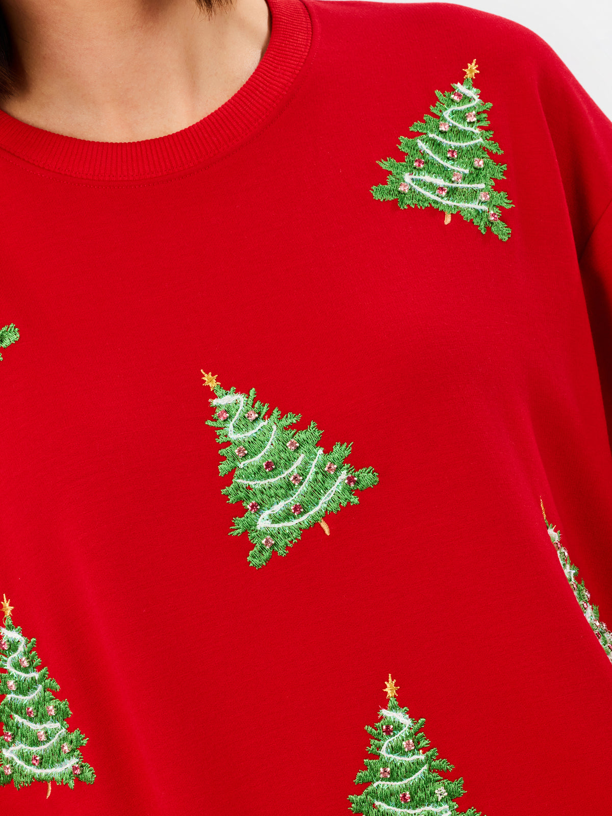 A close-up of a bright red crewneck sweatshirt featuring embroidered green Christmas trees with gold stars and white garlands.