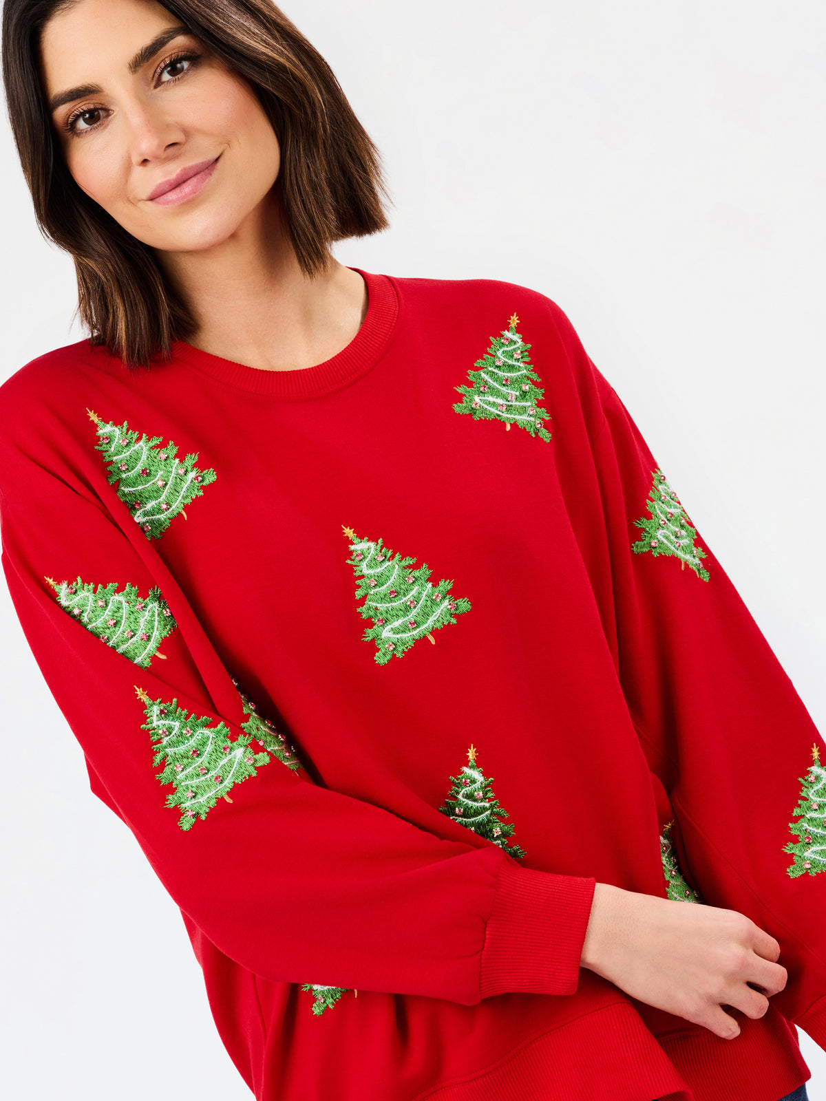 A woman wearing a bright red crewneck sweatshirt with green Christmas tree patterns, photographed against a clean white background.