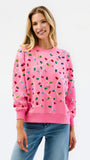 A woman wearing a pink crewneck sweatshirt decorated with colorful Christmas light bulbs, paired with blue jeans, against a plain white background.