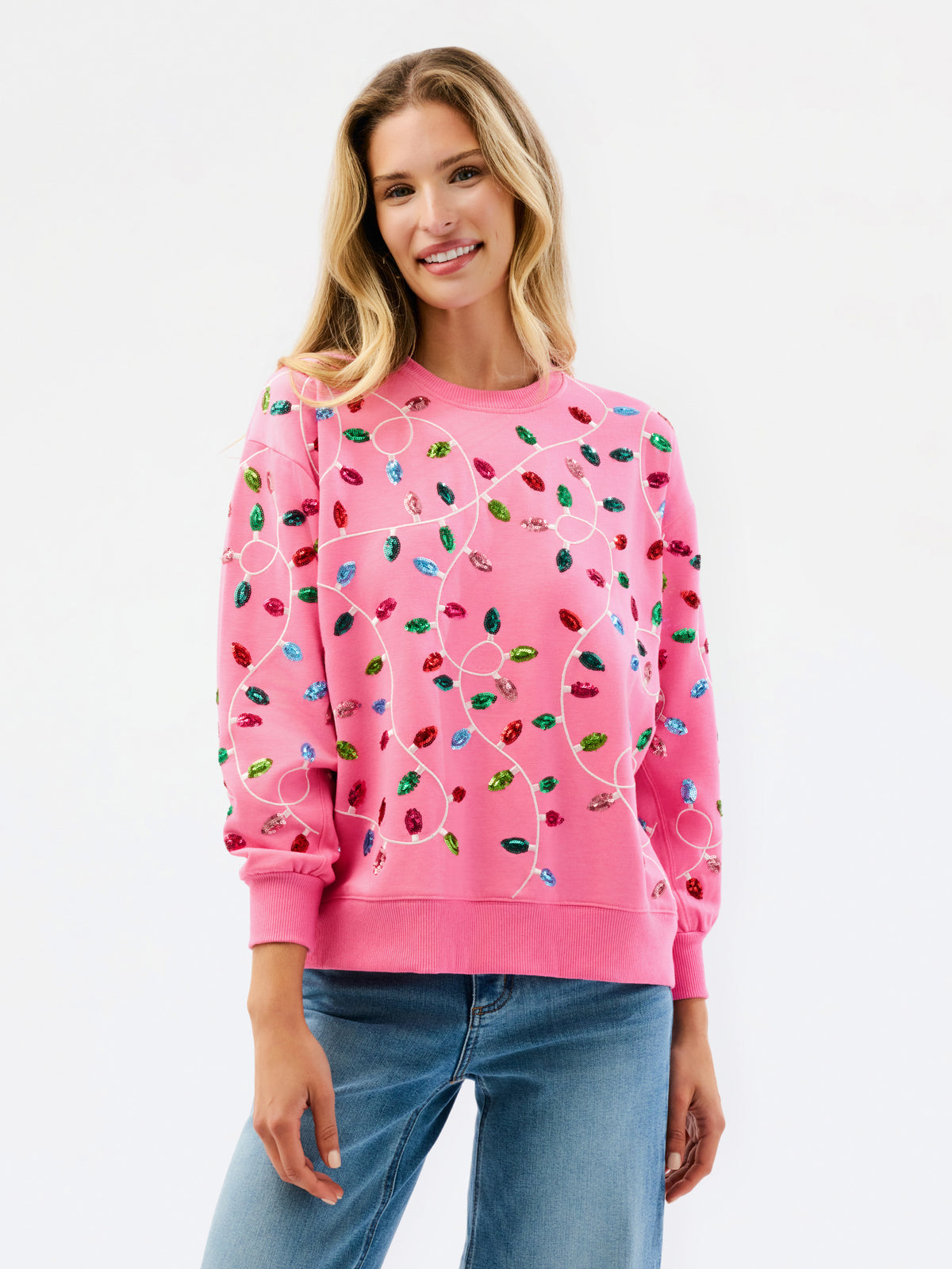 A woman wearing a pink crewneck sweatshirt decorated with colorful Christmas light bulbs, paired with blue jeans, against a plain white background.