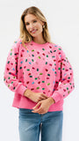 A woman wearing a pink crewneck sweatshirt decorated with colorful sequins that form a heart shape, paired with blue jeans, against a plain white background.