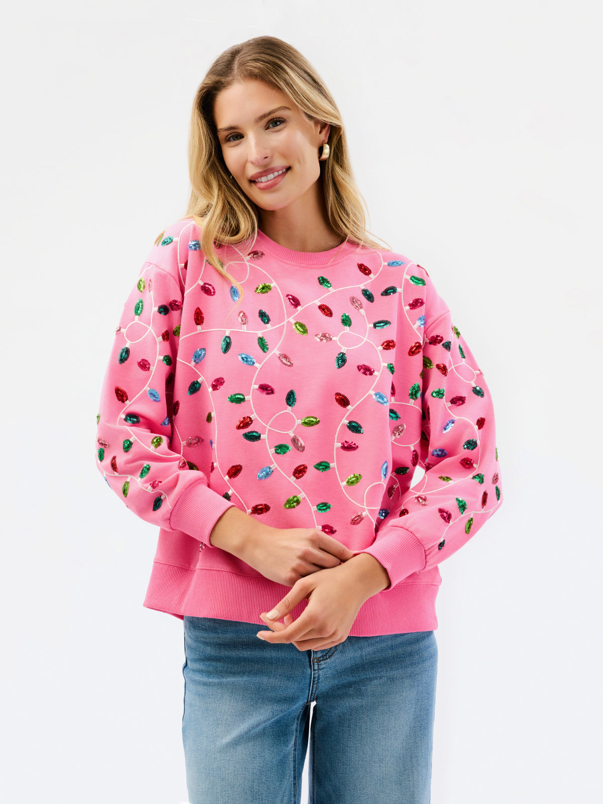 A woman wearing a pink crewneck sweatshirt decorated with colorful sequins that form a heart shape, paired with blue jeans, against a plain white background.