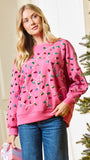 A woman wearing a pink crewneck sweatshirt decorated with colorful sequins that resemble Christmas lights, paired with blue jeans, standing next to a decorated Christmas tree.