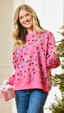 A woman wearing a pink crewneck sweatshirt decorated with colorful sequined Christmas light bulbs, standing next to a decorated Christmas tree and holding a pink gift box.
