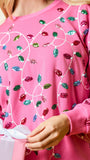 A close-up of a person wearing a bright pink crewneck sweatshirt decorated with sequined holiday light patterns in red, green, blue, and pink, holding a pink gift box with a white ribbon.