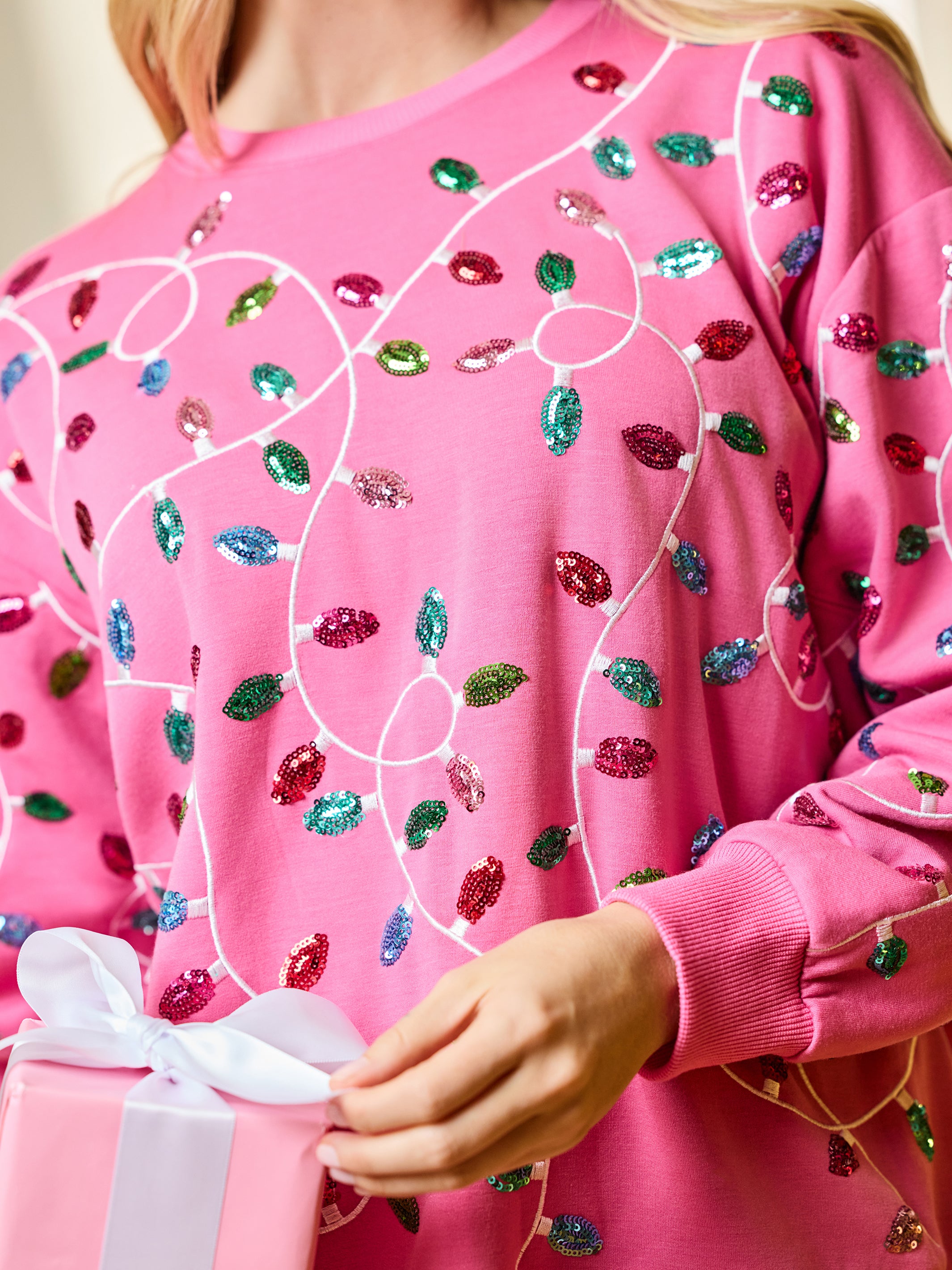 A close-up of a person wearing a bright pink crewneck sweatshirt decorated with sequined holiday light patterns in red, green, blue, and pink, holding a pink gift box with a white ribbon.