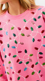 A close-up of a pink crewneck sweatshirt featuring an all-over pattern of colorful sequined Christmas light bulbs connected by white embroidered wire lines.