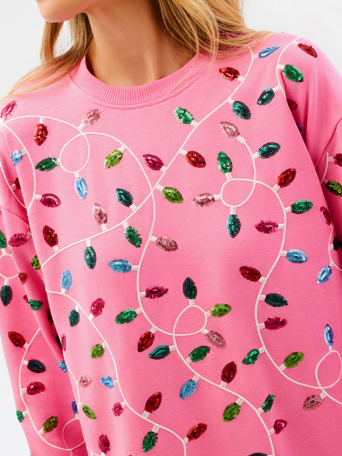 A close-up of a pink crewneck sweatshirt featuring an all-over pattern of colorful sequined Christmas light bulbs connected by white embroidered wire lines.