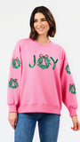 A woman wearing a pink crewneck sweatshirt with green sequined holiday wreath patches on the sleeves and 'JOY' spelled out in green sequins across the chest, paired with blue jeans against a plain white background.