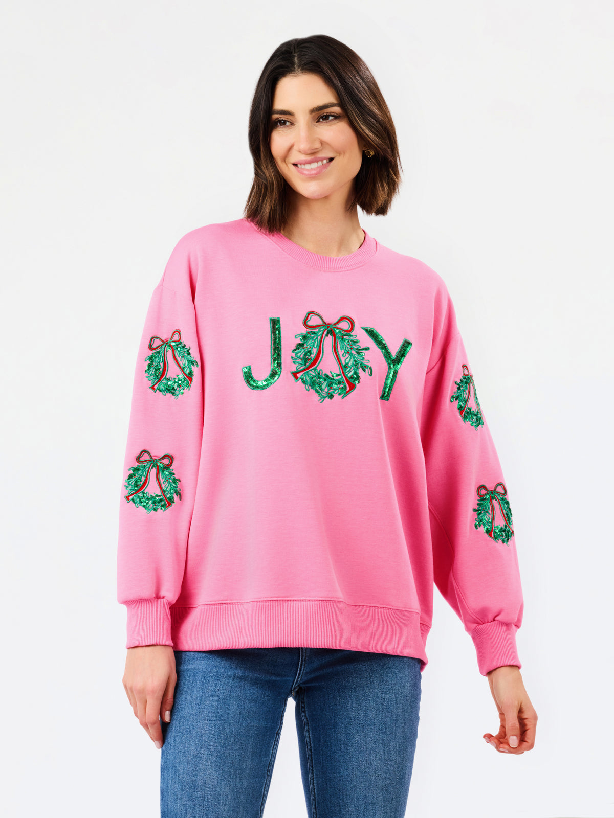 A woman wearing a pink crewneck sweatshirt with green sequined holiday wreath patches on the sleeves and 'JOY' spelled out in green sequins across the chest, paired with blue jeans against a plain white background.