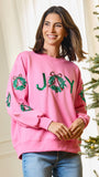 A woman wearing a pink crewneck sweatshirt with 'JOY' spelled out in green sequined wreaths, standing next to a lit Christmas tree.