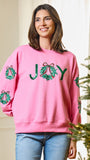 A woman wearing a pink crewneck sweatshirt with 'JOY' spelled out in green sequined wreaths on the front, standing next to a decorated Christmas tree.