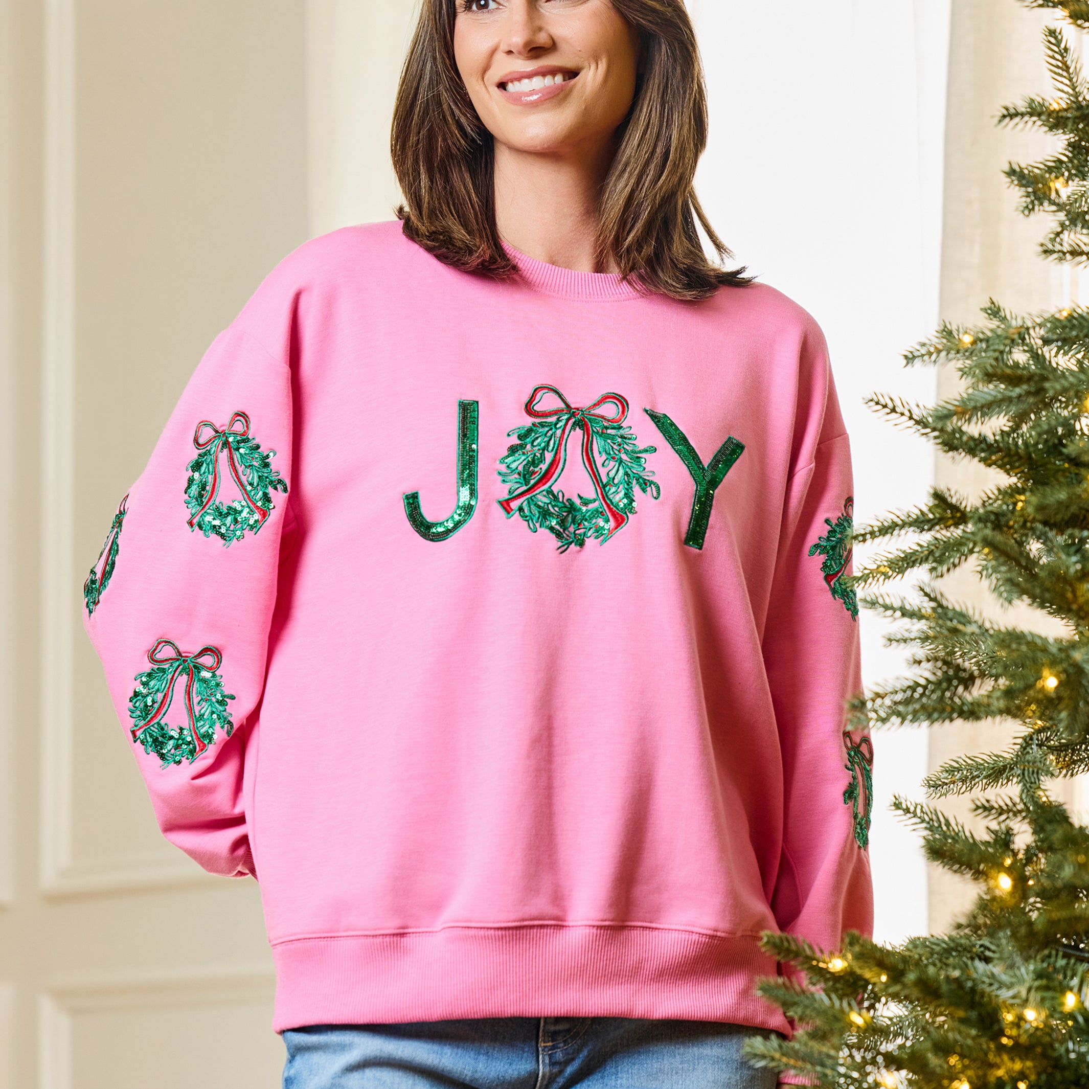 A woman wearing a pink crewneck sweatshirt with 'JOY' spelled out in green sequined wreaths on the front, standing next to a decorated Christmas tree.