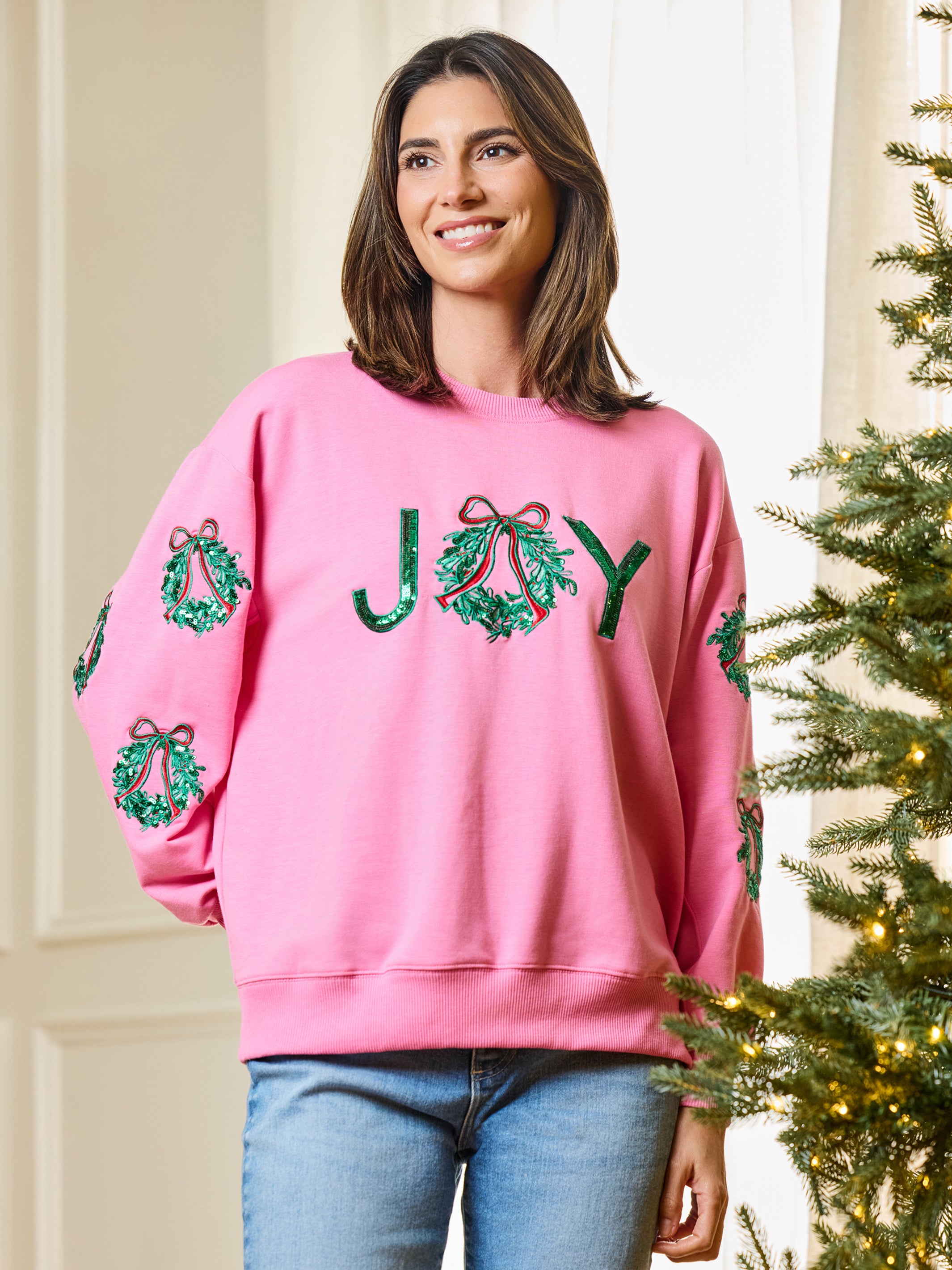 A woman wearing a pink crewneck sweatshirt with 'JOY' spelled out in green sequined wreaths on the front, standing next to a decorated Christmas tree.