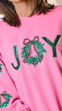 A close-up of a person wearing a bright pink crewneck sweatshirt with green sequined 'JOY' lettering and holly motifs on the front and sleeves.