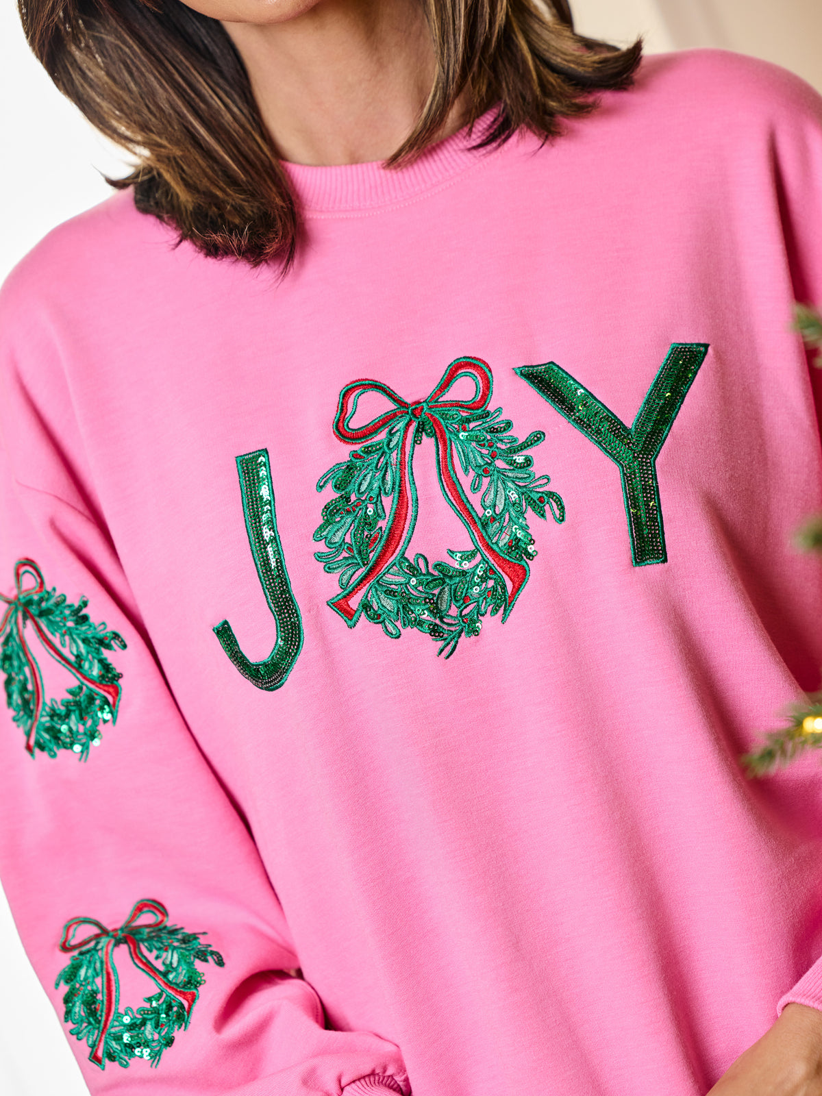 A close-up of a person wearing a bright pink crewneck sweatshirt with green sequined 'JOY' lettering and holly motifs on the front and sleeves.
