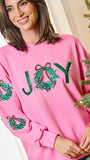 A woman wearing a pink crewneck sweatshirt with green sequined 'JOY' lettering and holly wreath motifs, posed next to a decorated Christmas tree.