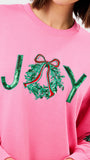A close-up of a pink crewneck sweatshirt featuring a sequined 'JOY' graphic on the chest, with the 'O' represented by a green sequin wreath with a red bow.
